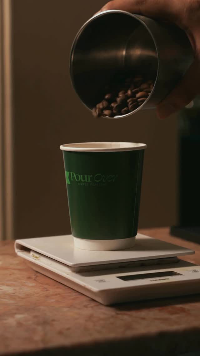 Hand pouring fresh-roasted beans into a Pour Over branded cup on a kitchen scale
